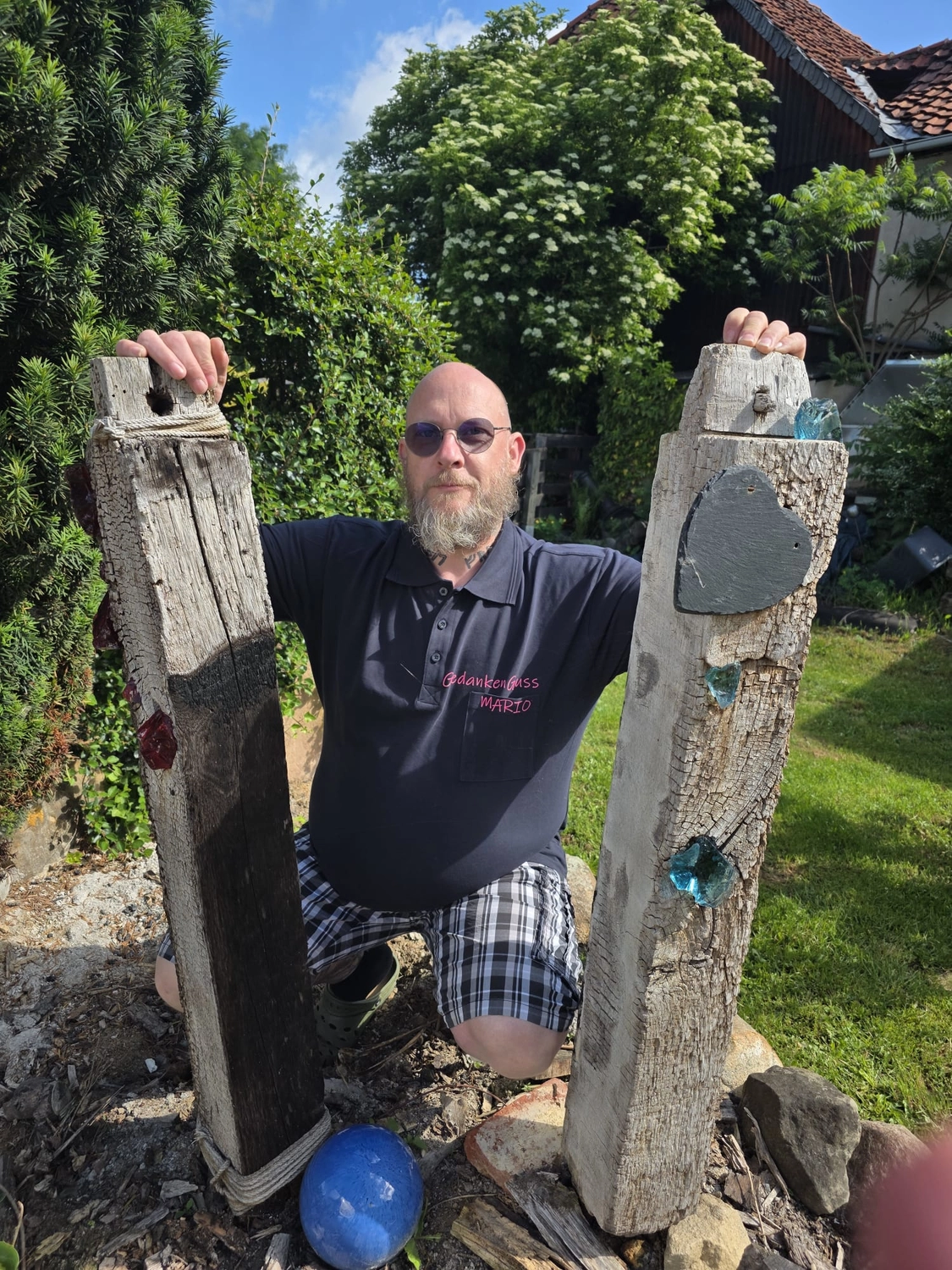 GedankenGuss - Holzskulpturen und abstrakten Kunstwerken - Mario Meinshausen mit Sonnenbrille steht draußen zwischen zwei großen Holzstämmen.