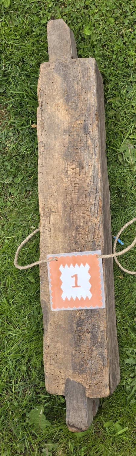GedankenGuss - Holzskulpturen und abstrakten Kunstwerken -  Eine kleine, weiß-orangefarbene Zierfahne mit einer Zahl hängt an einem dicken Seil über einem verwitterten Holzbrett, das auf einem Stück grünem Gras liegt.