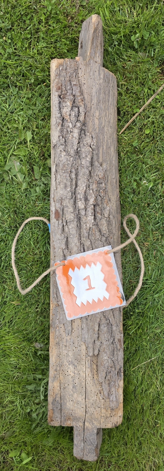 GedankenGuss - Holzskulpturen und abstrakten Kunstwerken -  Eine kleine, weiß-orangefarbene Zierfahne mit einer Zahl hängt an einem dicken Seil über einem verwitterten Holzbrett, das auf einem Stück grünem Gras liegt.