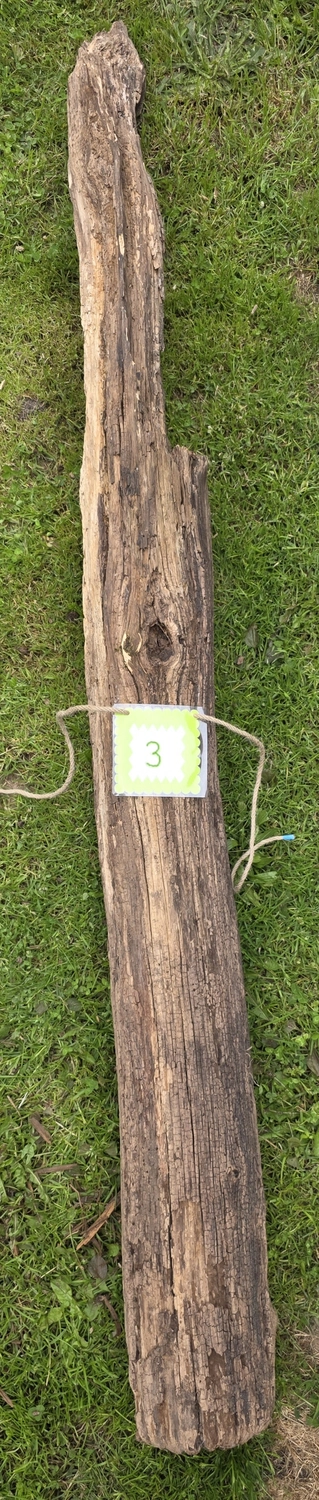 GedankenGuss - Holzskulpturen und abstrakten Kunstwerken -Eine kleine, hellgrüne und weiße Zierfahne mit einer Zahl hängt an einem dicken Seil über einem verwitterten Holzbrett, das auf einem Stück grünem Gras liegt.