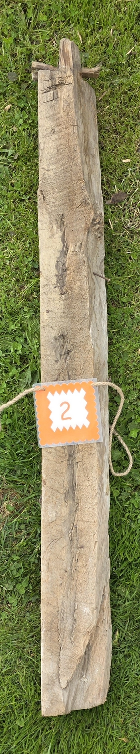 GedankenGuss - Holzskulpturen und abstrakten Kunstwerken -  Eine kleine, weiß-orangefarbene Zierfahne mit einer Zahl hängt an einem dicken Seil über einem verwitterten Holzbrett, das auf einem Stück grünem Gras liegt.