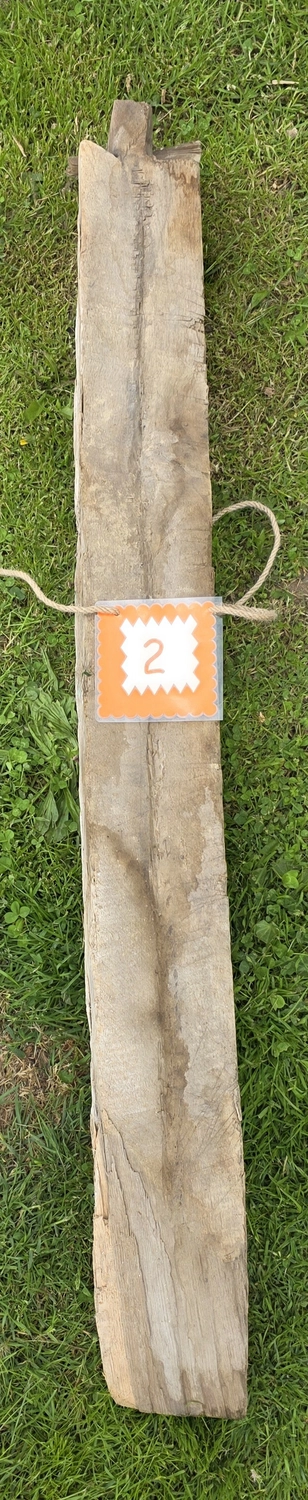 GedankenGuss - Holzskulpturen und abstrakten Kunstwerken -  Eine kleine, weiß-orangefarbene Zierfahne mit einer Zahl hängt an einem dicken Seil über einem verwitterten Holzbrett, das auf einem Stück grünem Gras liegt.