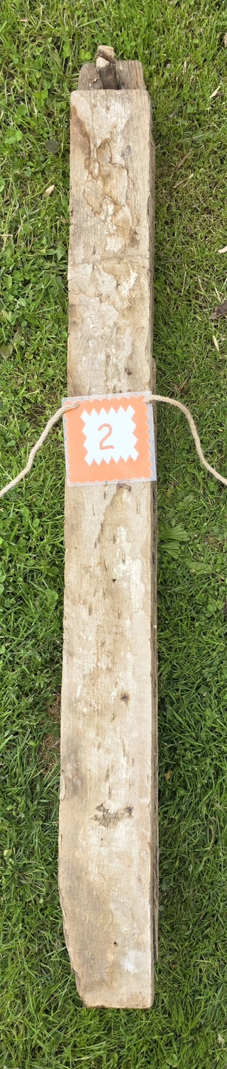 GedankenGuss - Holzskulpturen und abstrakten Kunstwerken -  Eine kleine, weiß-orangefarbene Zierfahne mit einer Zahl hängt an einem dicken Seil über einem verwitterten Holzbrett, das auf einem Stück grünem Gras liegt.