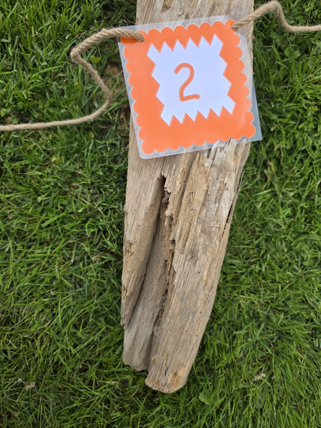 GedankenGuss - Holzskulpturen und abstrakten Kunstwerken -  Eine kleine, weiß-orangefarbene Zierfahne mit einer Zahl hängt an einem dicken Seil über einem verwitterten Holzbrett, das auf einem Stück grünem Gras liegt.