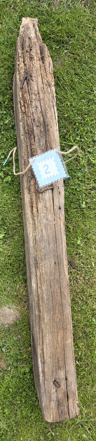 GedankenGuss - Holzskulpturen und abstrakten Kunstwerken -Eine kleine Blau und weiße Zierfahne mit einer Zahl hängt an einem dicken Seil über einem verwitterten Holzbrett, das auf einem Stück grünem Gras liegt.