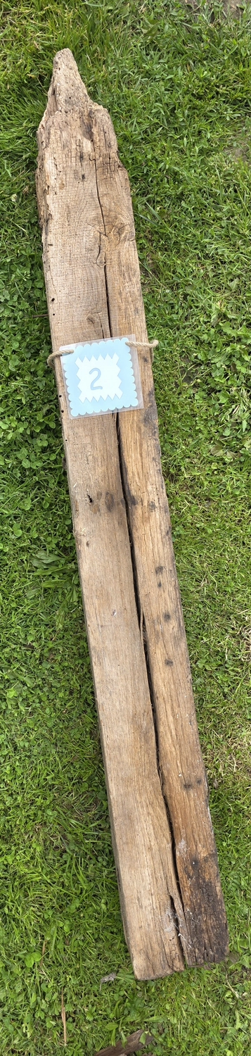 GedankenGuss - Holzskulpturen und abstrakten Kunstwerken -Eine kleine Blau und weiße Zierfahne mit einer Zahl hängt an einem dicken Seil über einem verwitterten Holzbrett, das auf einem Stück grünem Gras liegt.