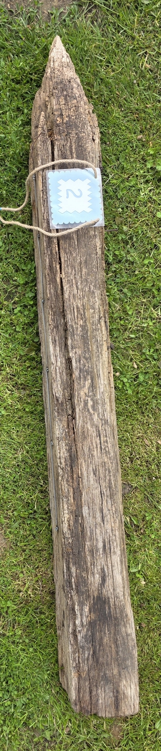GedankenGuss - Holzskulpturen und abstrakten Kunstwerken -Eine kleine Blau und weiße Zierfahne mit einer Zahl hängt an einem dicken Seil über einem verwitterten Holzbrett, das auf einem Stück grünem Gras liegt.