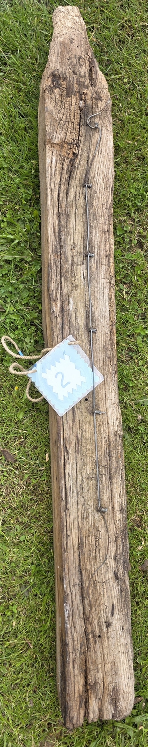 GedankenGuss - Holzskulpturen und abstrakten Kunstwerken -Eine kleine Blau und weiße Zierfahne mit einer Zahl hängt an einem dicken Seil über einem verwitterten Holzbrett, das auf einem Stück grünem Gras liegt.