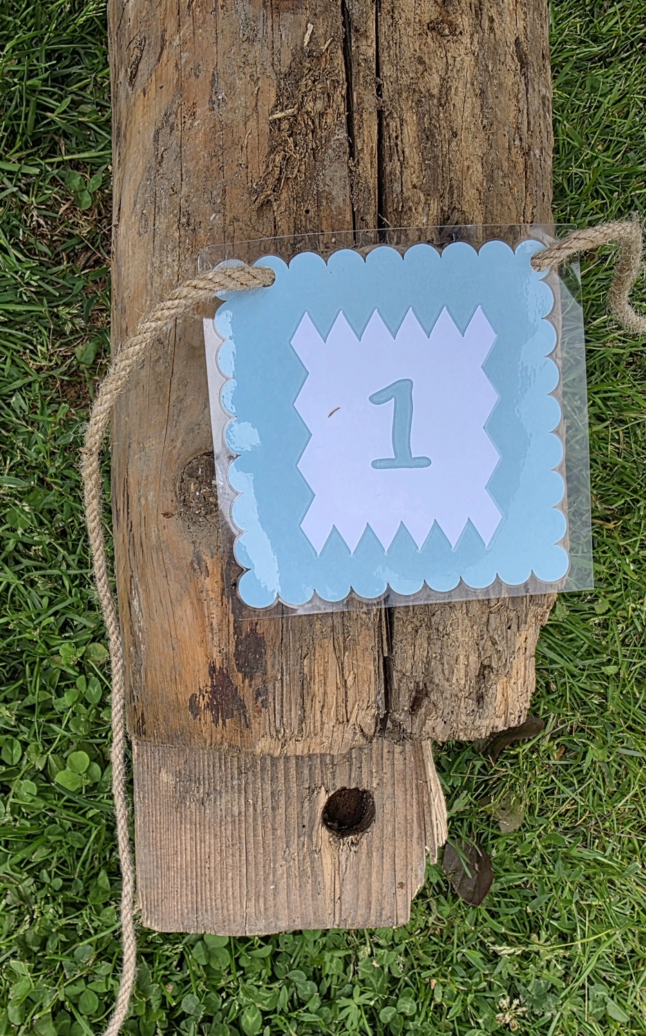 GedankenGuss - Holzskulpturen und abstrakten Kunstwerken -Eine kleine Blau und weiße Zierfahne mit einer Zahl hängt an einem dicken Seil über einem verwitterten Holzbrett, das auf einem Stück grünem Gras liegt.