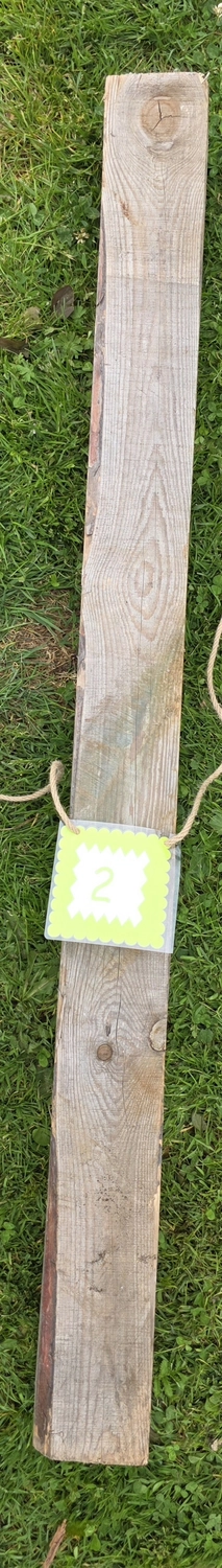 GedankenGuss - Holzskulpturen und abstrakten Kunstwerken -Eine kleine, hellgrüne und weiße Zierfahne mit einer Zahl hängt an einem dicken Seil über einem verwitterten Holzbrett, das auf einem Stück grünem Gras liegt.
