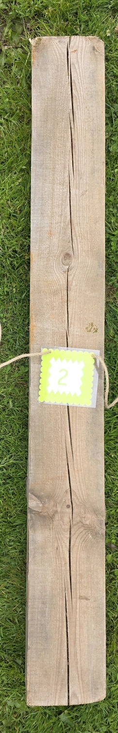 GedankenGuss - Holzskulpturen und abstrakten Kunstwerken -Eine kleine, hellgrüne und weiße Zierfahne mit einer Zahl hängt an einem dicken Seil über einem verwitterten Holzbrett, das auf einem Stück grünem Gras liegt.