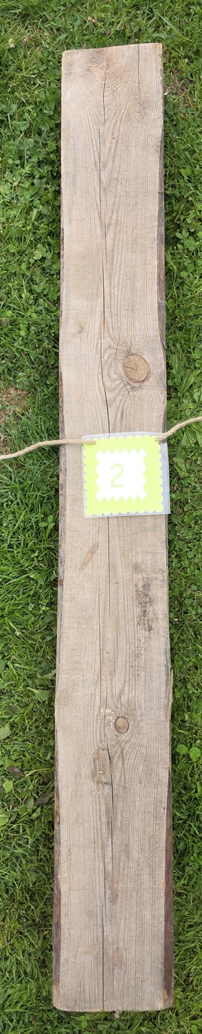 GedankenGuss - Holzskulpturen und abstrakten Kunstwerken -Eine kleine, hellgrüne und weiße Zierfahne mit einer Zahl hängt an einem dicken Seil über einem verwitterten Holzbrett, das auf einem Stück grünem Gras liegt.