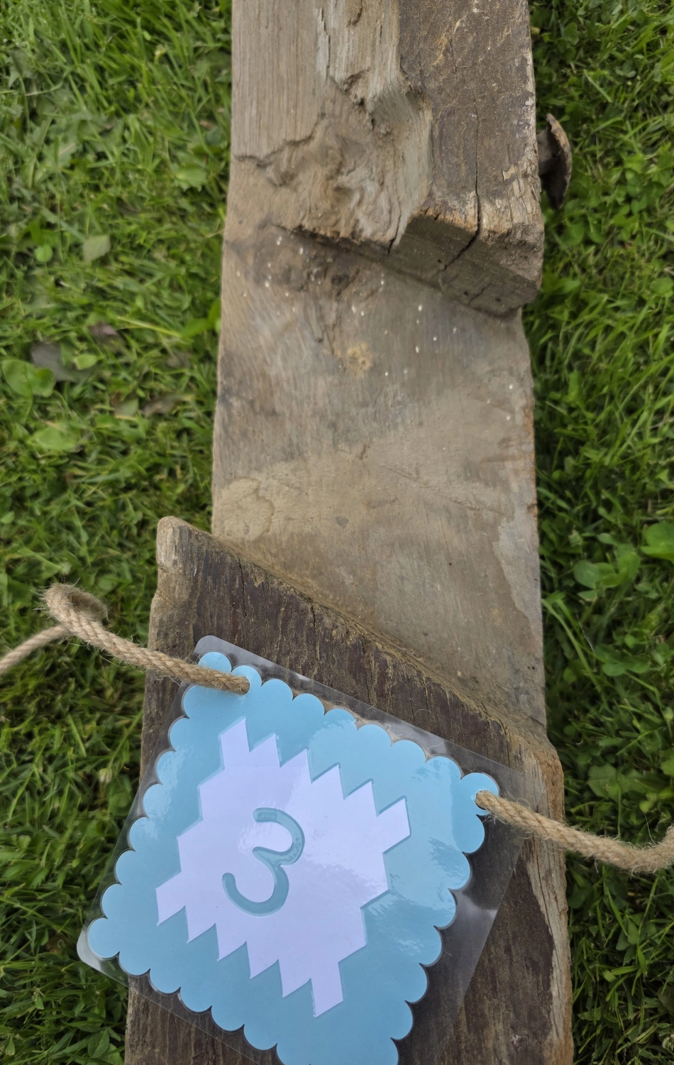 GedankenGuss - Holzskulpturen und abstrakten Kunstwerken -Eine kleine Blau und weiße Zierfahne mit einer Zahl hängt an einem dicken Seil über einem verwitterten Holzbrett, das auf einem Stück grünem Gras liegt.