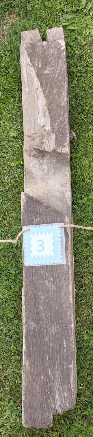 GedankenGuss - Holzskulpturen und abstrakten Kunstwerken -Eine kleine Blau und weiße Zierfahne mit einer Zahl hängt an einem dicken Seil über einem verwitterten Holzbrett, das auf einem Stück grünem Gras liegt.