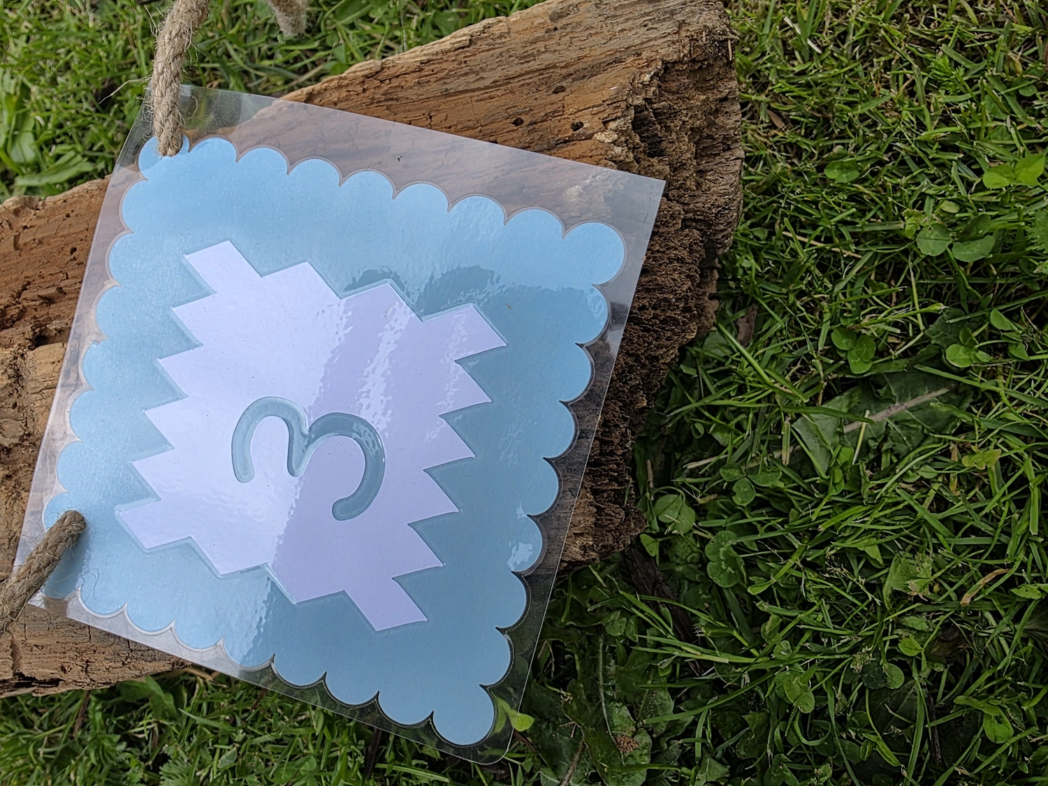 GedankenGuss - Holzskulpturen und abstrakten Kunstwerken -Eine kleine Blau und weiße Zierfahne mit einer Zahl hängt an einem dicken Seil über einem verwitterten Holzbrett, das auf einem Stück grünem Gras liegt.