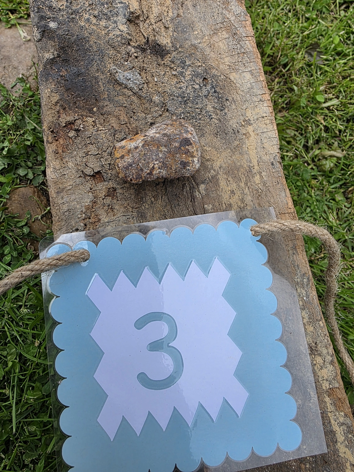 GedankenGuss - Holzskulpturen und abstrakten Kunstwerken -Eine kleine Blau und weiße Zierfahne mit einer Zahl hängt an einem dicken Seil über einem verwitterten Holzbrett, das auf einem Stück grünem Gras liegt.