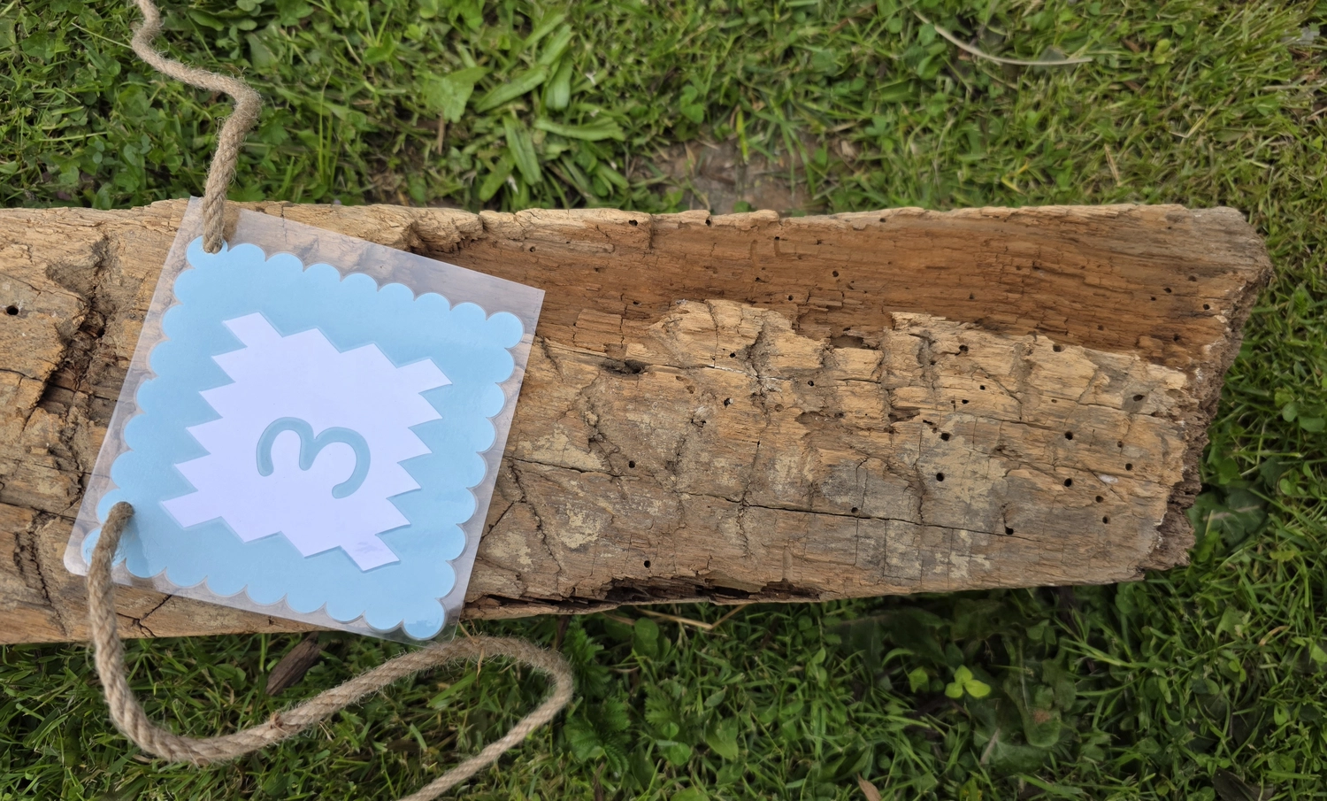 GedankenGuss - Holzskulpturen und abstrakten Kunstwerken -Eine kleine Blau und weiße Zierfahne mit einer Zahl hängt an einem dicken Seil über einem verwitterten Holzbrett, das auf einem Stück grünem Gras liegt.
