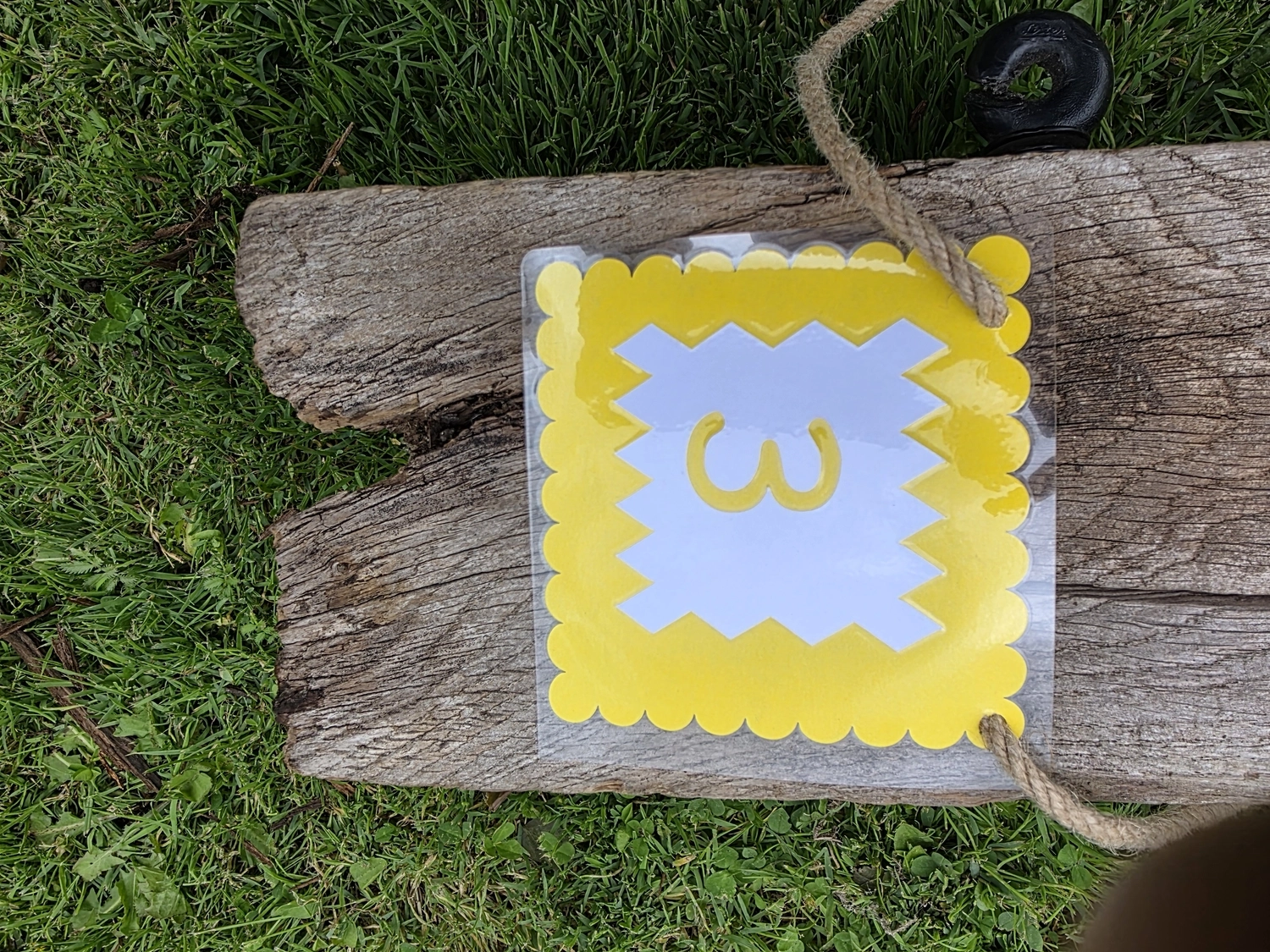 GedankenGuss - Holzskulpturen und abstrakten Kunstwerken - Eine kleine, gelb-weiße Zierfahne mit der Nummer hängt an einem dicken Seil über einem verwitterten Holzbrett, das auf einem Stück grünem Gras liegt.