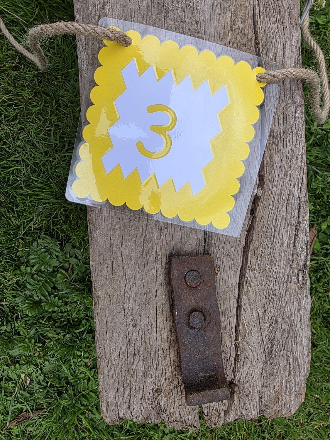 GedankenGuss - Holzskulpturen und abstrakten Kunstwerken - Eine kleine, gelb-weiße Zierfahne mit der Nummer hängt an einem dicken Seil über einem verwitterten Holzbrett, das auf einem Stück grünem Gras liegt.