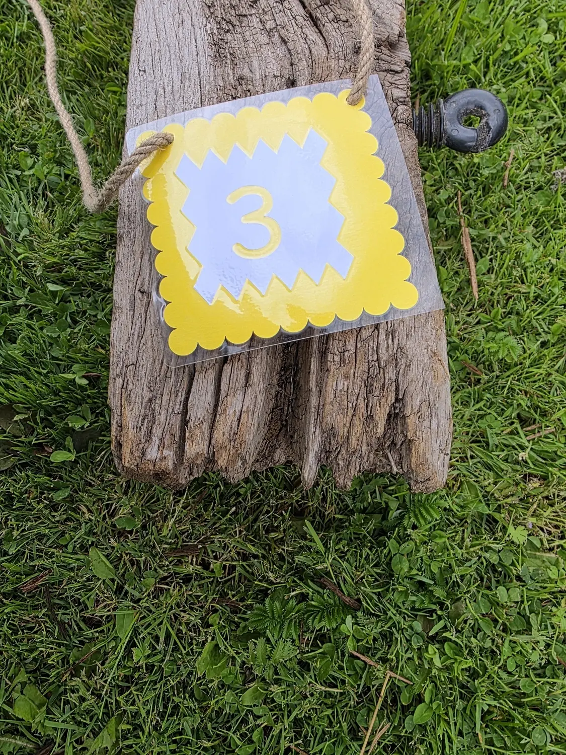 GedankenGuss - Holzskulpturen und abstrakten Kunstwerken - Eine kleine, gelb-weiße Zierfahne mit der Nummer hängt an einem dicken Seil über einem verwitterten Holzbrett, das auf einem Stück grünem Gras liegt.