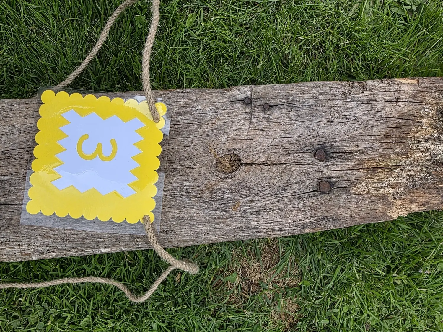GedankenGuss - Holzskulpturen und abstrakten Kunstwerken - Eine kleine, gelb-weiße Zierfahne mit der Nummer hängt an einem dicken Seil über einem verwitterten Holzbrett, das auf einem Stück grünem Gras liegt.