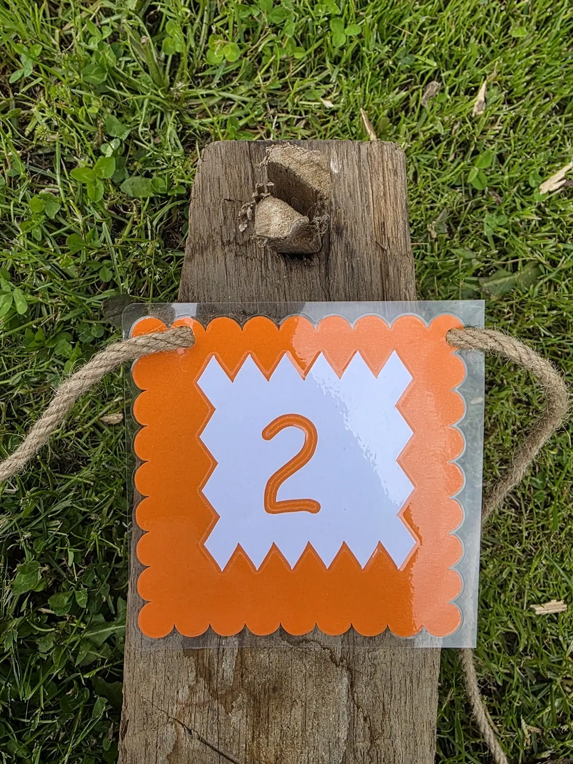 GedankenGuss - Holzskulpturen und abstrakten Kunstwerken -  Eine kleine, weiß-orangefarbene Zierfahne mit einer Zahl hängt an einem dicken Seil über einem verwitterten Holzbrett, das auf einem Stück grünem Gras liegt.