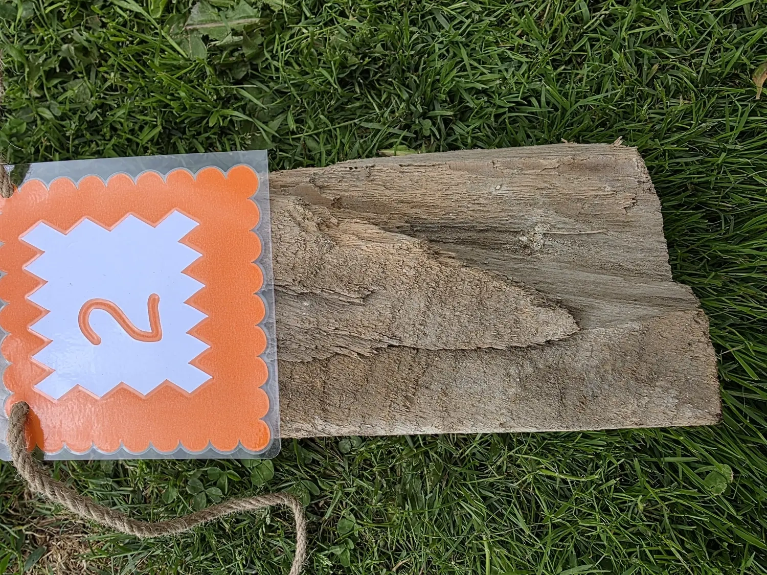 GedankenGuss - Holzskulpturen und abstrakten Kunstwerken -  Eine kleine, weiß-orangefarbene Zierfahne mit einer Zahl hängt an einem dicken Seil über einem verwitterten Holzbrett, das auf einem Stück grünem Gras liegt.