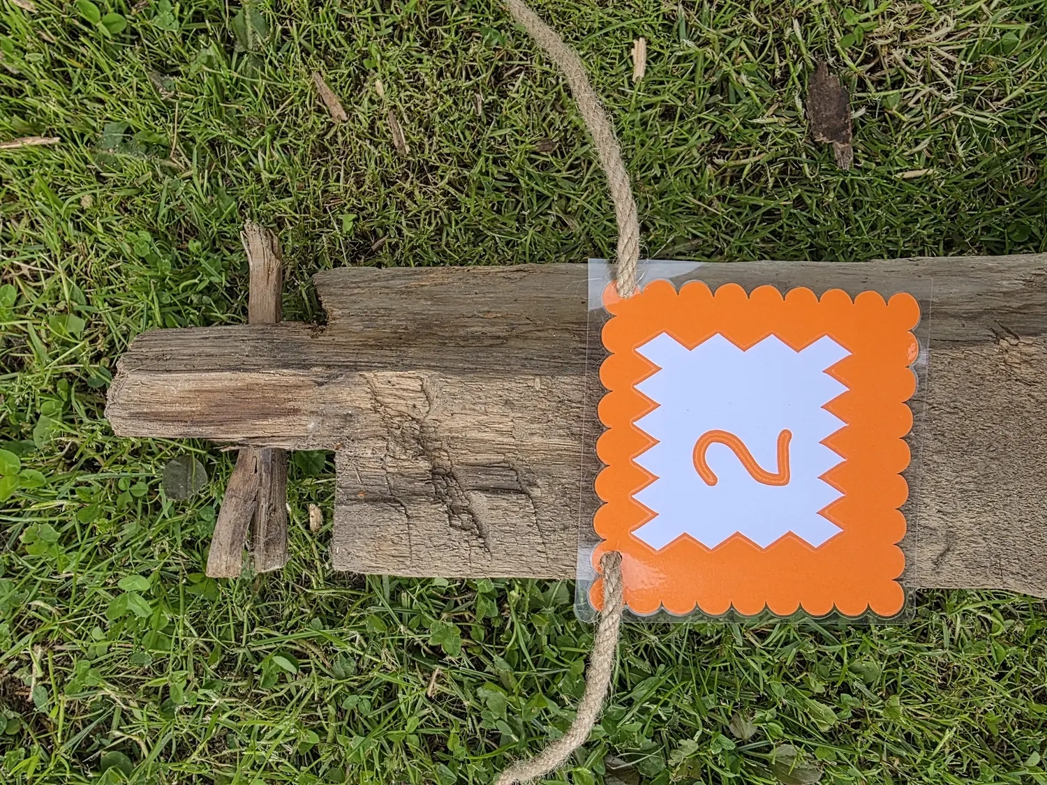 GedankenGuss - Holzskulpturen und abstrakten Kunstwerken -  Eine kleine, weiß-orangefarbene Zierfahne mit einer Zahl hängt an einem dicken Seil über einem verwitterten Holzbrett, das auf einem Stück grünem Gras liegt.