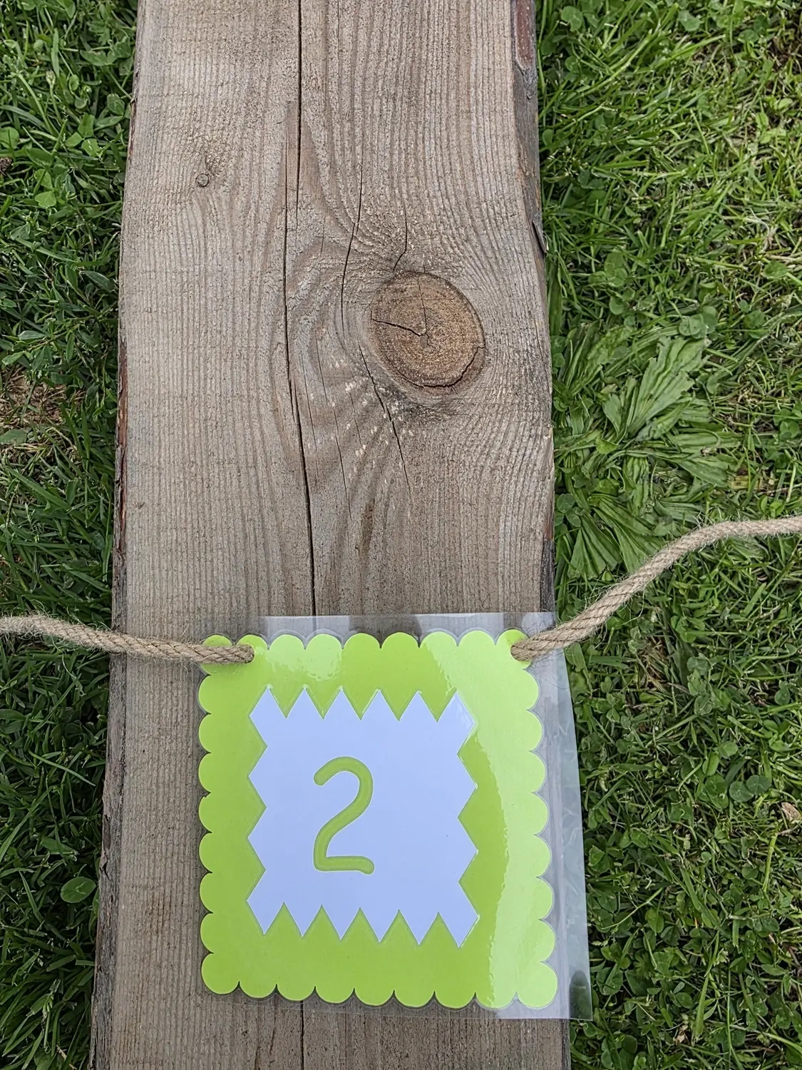 GedankenGuss - Holzskulpturen und abstrakten Kunstwerken -Eine kleine, hellgrüne und weiße Zierfahne mit einer Zahl hängt an einem dicken Seil über einem verwitterten Holzbrett, das auf einem Stück grünem Gras liegt.