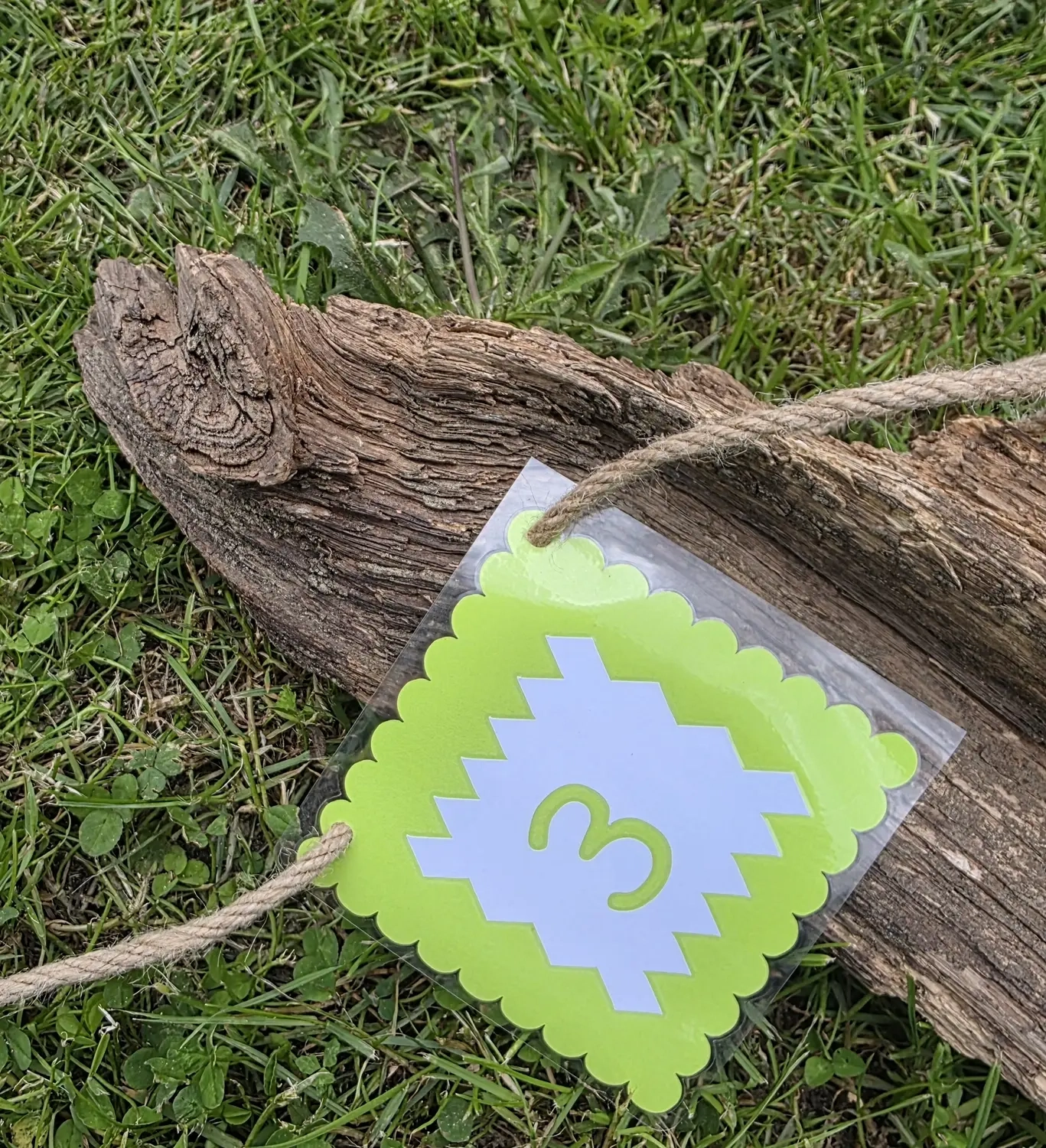 GedankenGuss - Holzskulpturen und abstrakten Kunstwerken -Eine kleine, hellgrüne und weiße Zierfahne mit einer Zahl hängt an einem dicken Seil über einem verwitterten Holzbrett, das auf einem Stück grünem Gras liegt.