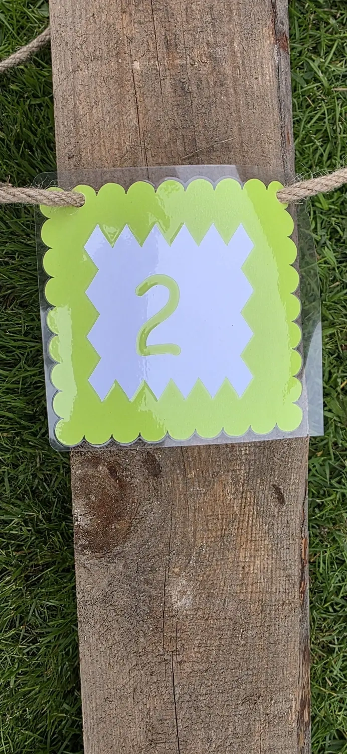 GedankenGuss - Holzskulpturen und abstrakten Kunstwerken -Eine kleine, hellgrüne und weiße Zierfahne mit einer Zahl hängt an einem dicken Seil über einem verwitterten Holzbrett, das auf einem Stück grünem Gras liegt.