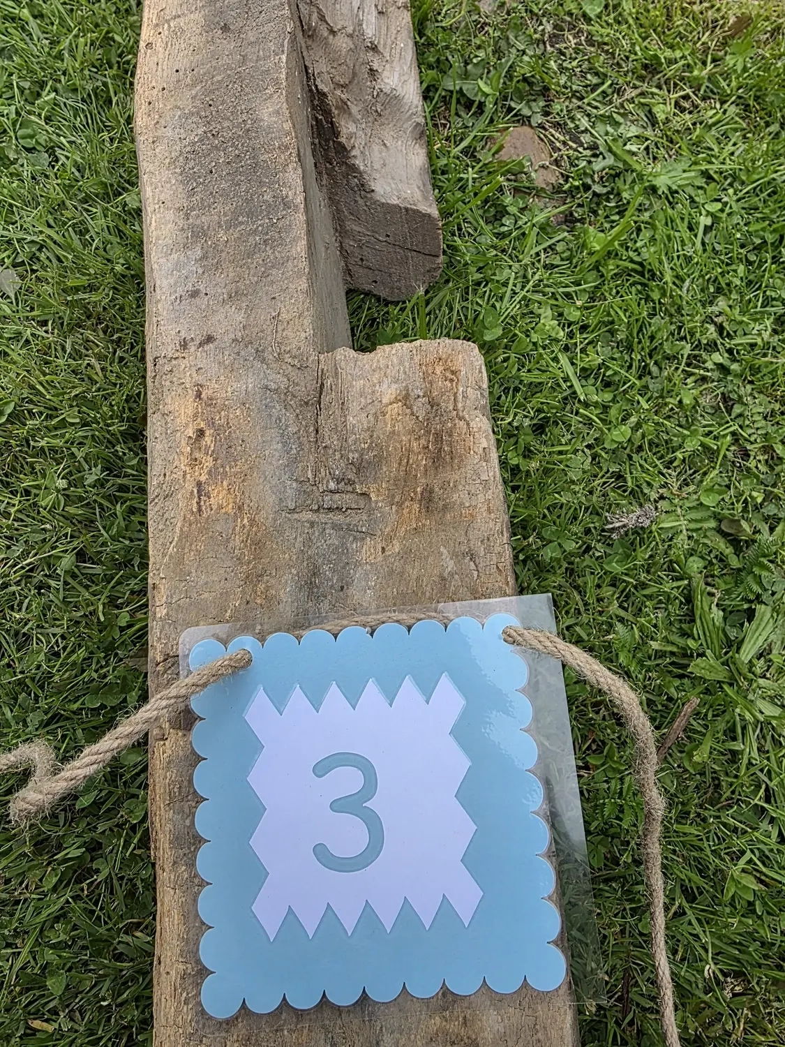 GedankenGuss - Holzskulpturen und abstrakten Kunstwerken -Eine kleine Blau und weiße Zierfahne mit einer Zahl hängt an einem dicken Seil über einem verwitterten Holzbrett, das auf einem Stück grünem Gras liegt.