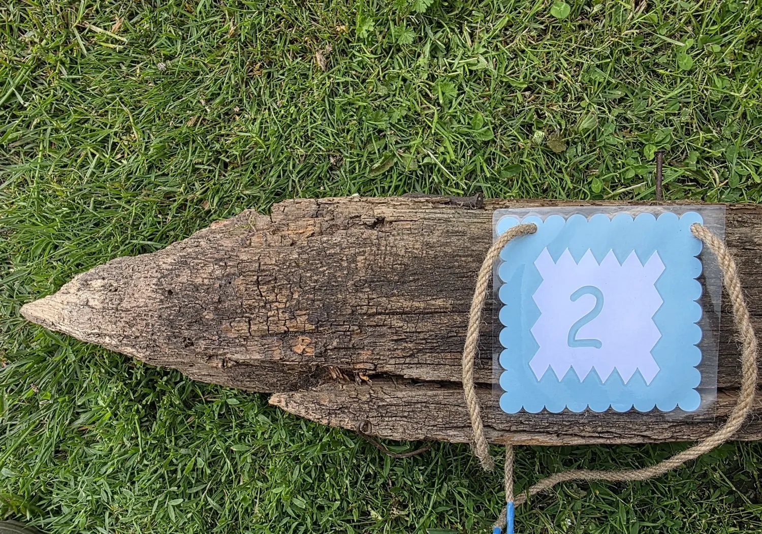 GedankenGuss - Holzskulpturen und abstrakten Kunstwerken -Eine kleine Blau und weiße Zierfahne mit einer Zahl hängt an einem dicken Seil über einem verwitterten Holzbrett, das auf einem Stück grünem Gras liegt.