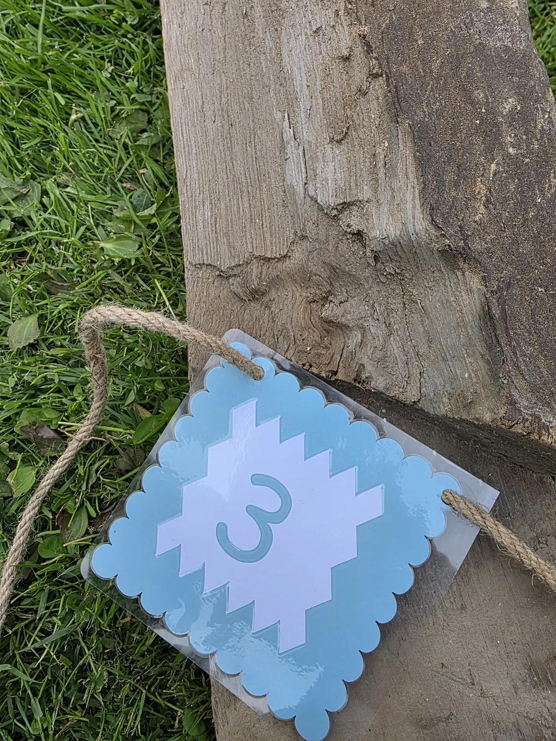 GedankenGuss - Holzskulpturen und abstrakten Kunstwerken -Eine kleine Blau und weiße Zierfahne mit einer Zahl hängt an einem dicken Seil über einem verwitterten Holzbrett, das auf einem Stück grünem Gras liegt.