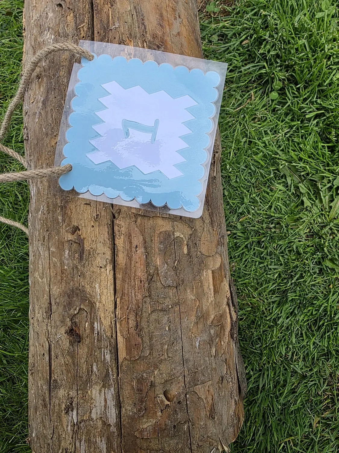 GedankenGuss - Holzskulpturen und abstrakten Kunstwerken -Eine kleine Blau und weiße Zierfahne mit einer Zahl hängt an einem dicken Seil über einem verwitterten Holzbrett, das auf einem Stück grünem Gras liegt.