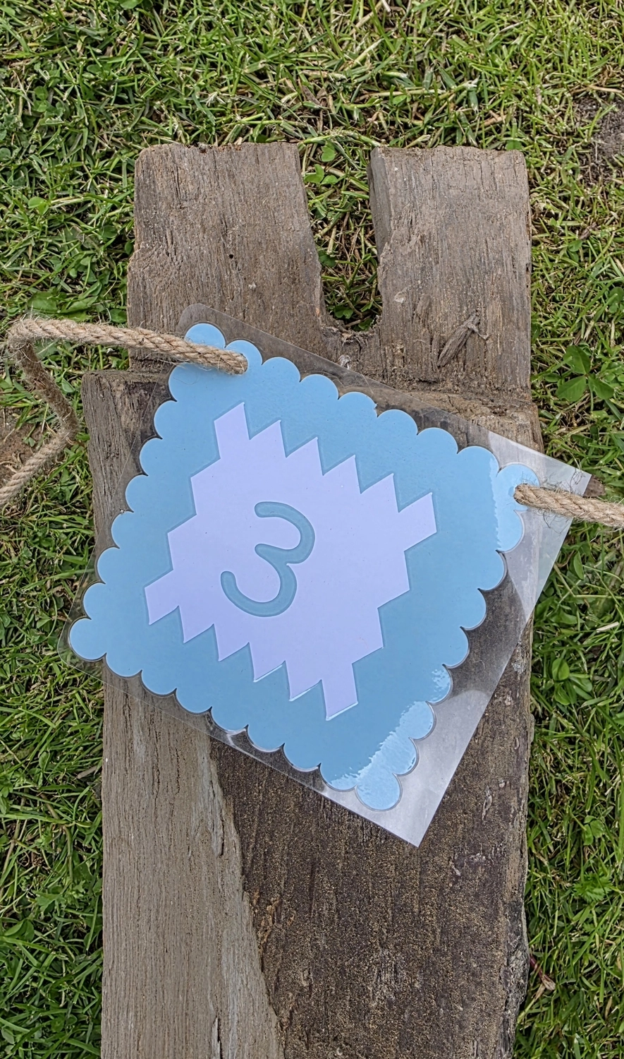 GedankenGuss - Holzskulpturen und abstrakten Kunstwerken -Eine kleine Blau und weiße Zierfahne mit einer Zahl hängt an einem dicken Seil über einem verwitterten Holzbrett, das auf einem Stück grünem Gras liegt.