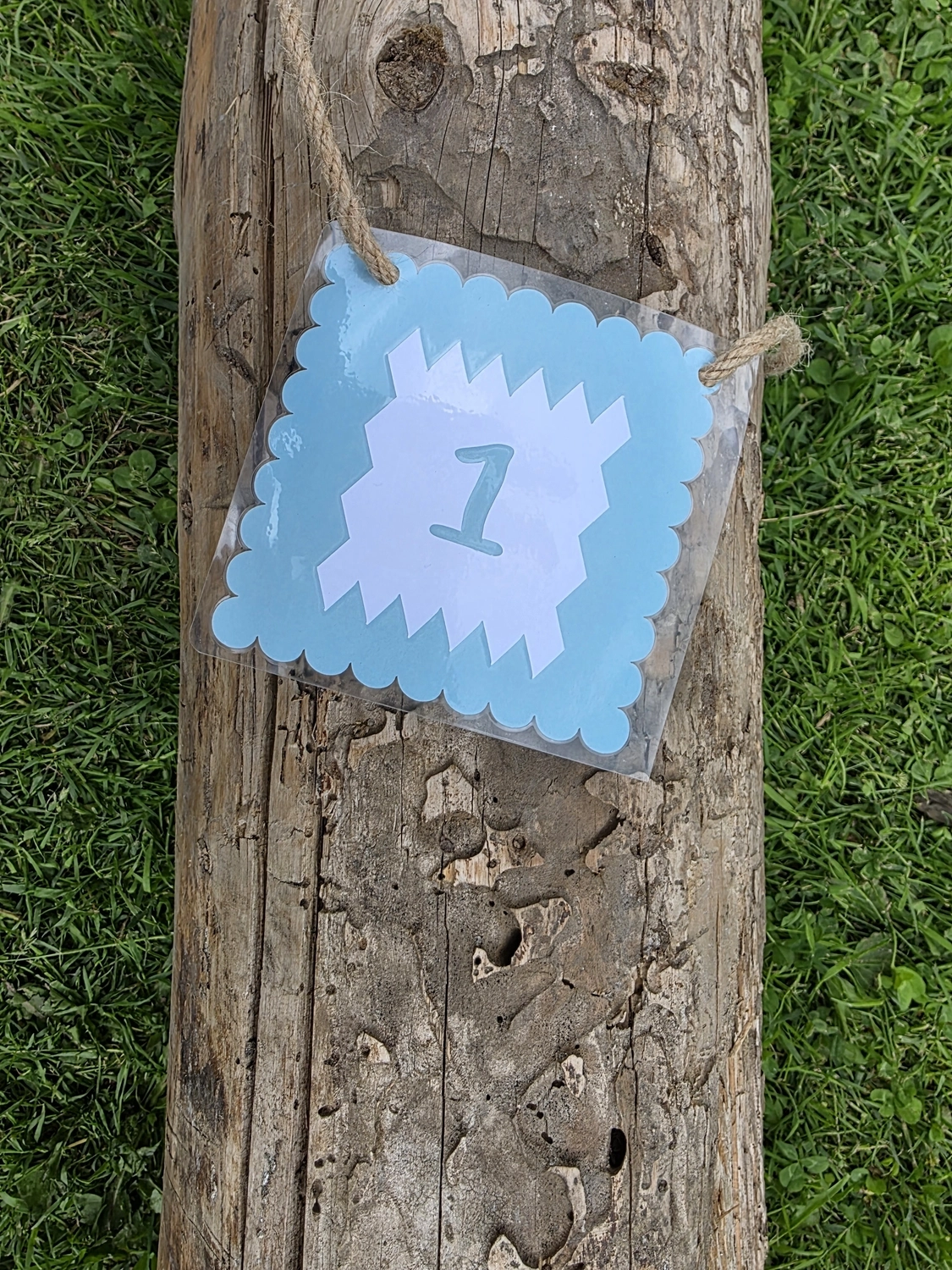 GedankenGuss - Holzskulpturen und abstrakten Kunstwerken -Eine kleine Blau und weiße Zierfahne mit einer Zahl hängt an einem dicken Seil über einem verwitterten Holzbrett, das auf einem Stück grünem Gras liegt.