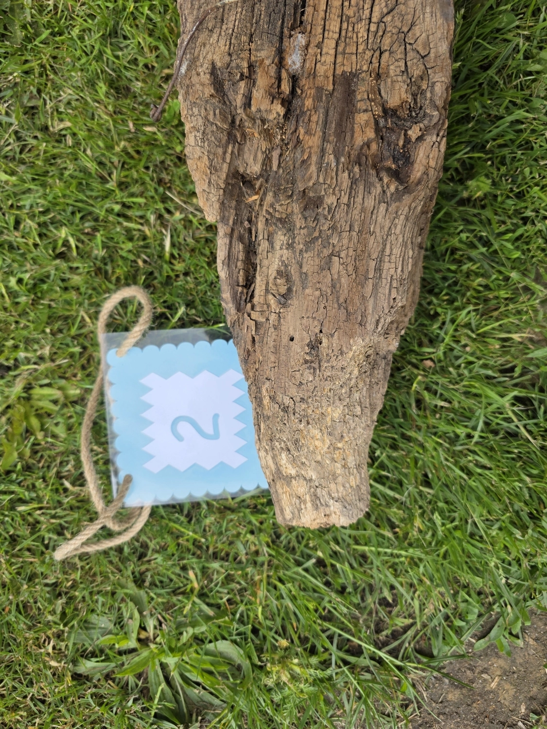 GedankenGuss - Holzskulpturen und abstrakten Kunstwerken -Eine kleine Blau und weiße Zierfahne mit einer Zahl hängt an einem dicken Seil über einem verwitterten Holzbrett, das auf einem Stück grünem Gras liegt.